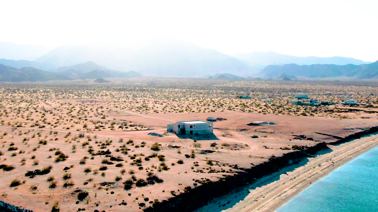 Drone-style frame over Bahia De Los Angeles with bright turquoise water at the right edge, a narrow exposed shore, shrub-dotted desert flats, several small structures, distant hazy mountains, and no visible people.