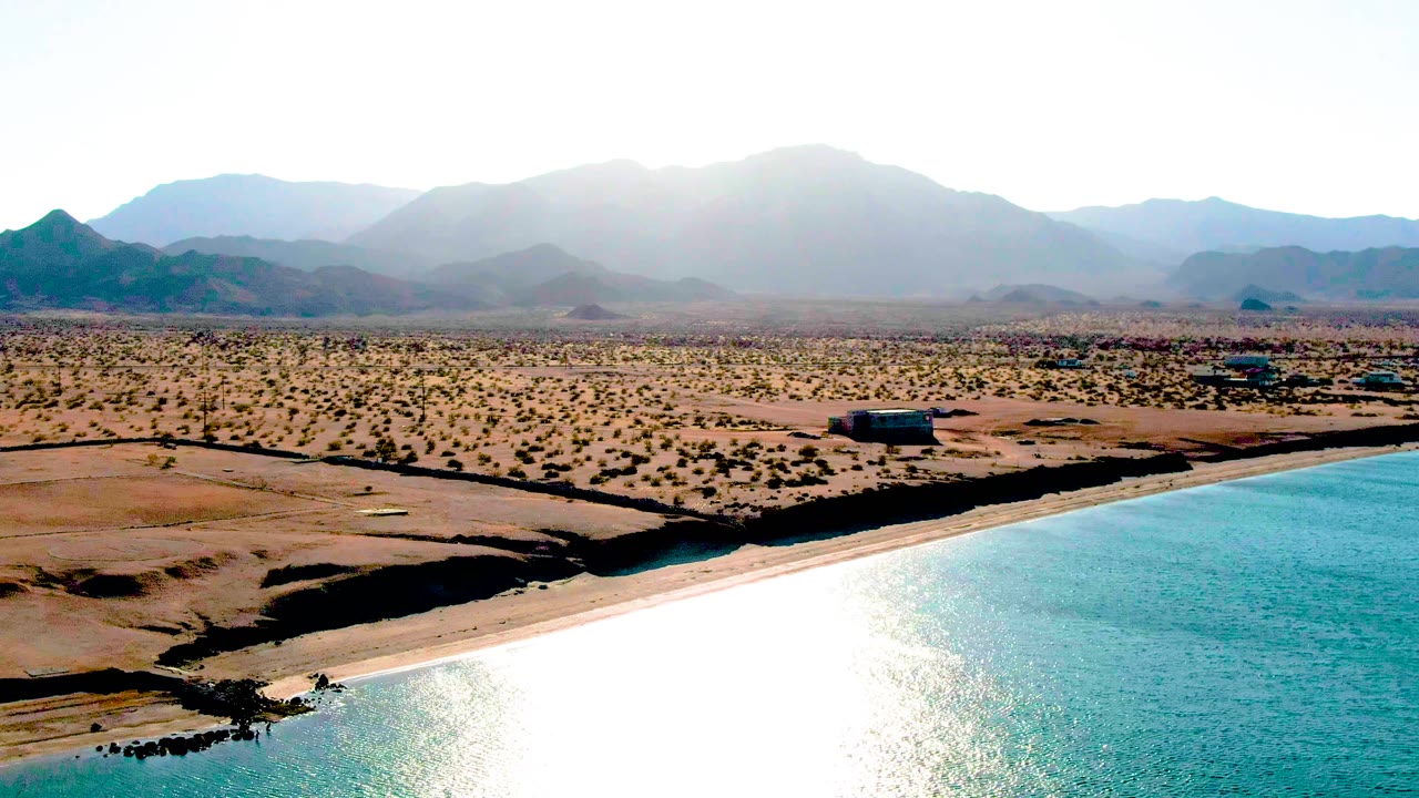 Wide aerial view of Bahia De Los Angeles showing turquoise water, bright sun glare, sandy desert shoreline, sparse scrub, low buildings, and hazy mountains, with no visible people.