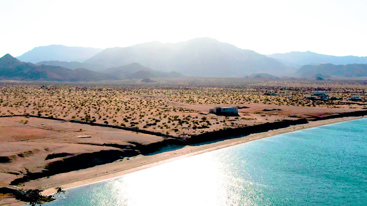 Bahia De Los Angeles appears from above with a strong white reflection on rippled turquoise water, a narrow sandy shore, dry patterned terrain, small buildings, and hazy mountains, with no visible people.