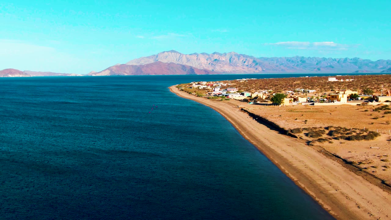 Opening aerial frame of Bahia De Los Angeles with turquoise ocean on the left, a curved sandy beach on the right, village buildings beyond the sand, distant mountains, and no visible people.