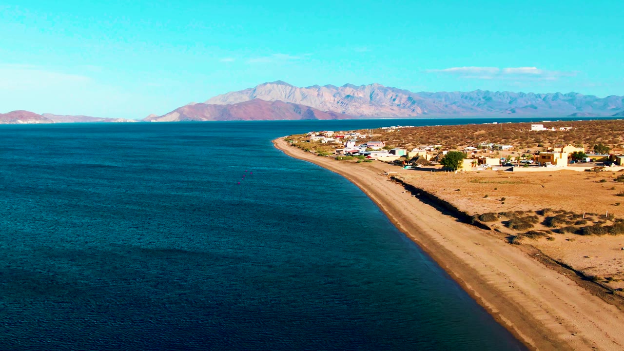 At 0:01, Bahia De Los Angeles appears from above with blue water filling the left side, the beach forming a diagonal shoreline, low village buildings on desert ground, mountains in the distance, and no visible people.