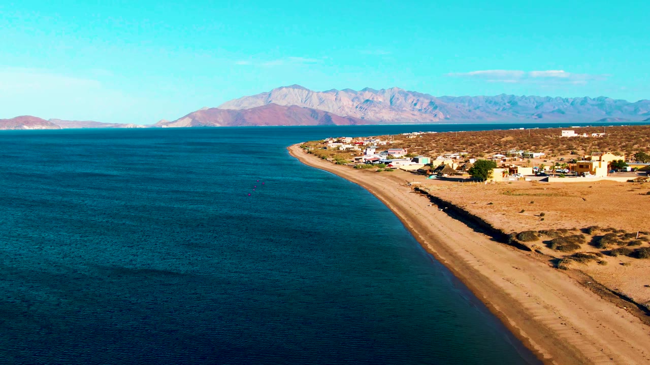 At 0:02, the aerial composition shows a wider stretch of Bahia De Los Angeles water, the sandy beach tapering toward the village, dry terrain behind the buildings, distant mountains, and no visible people.