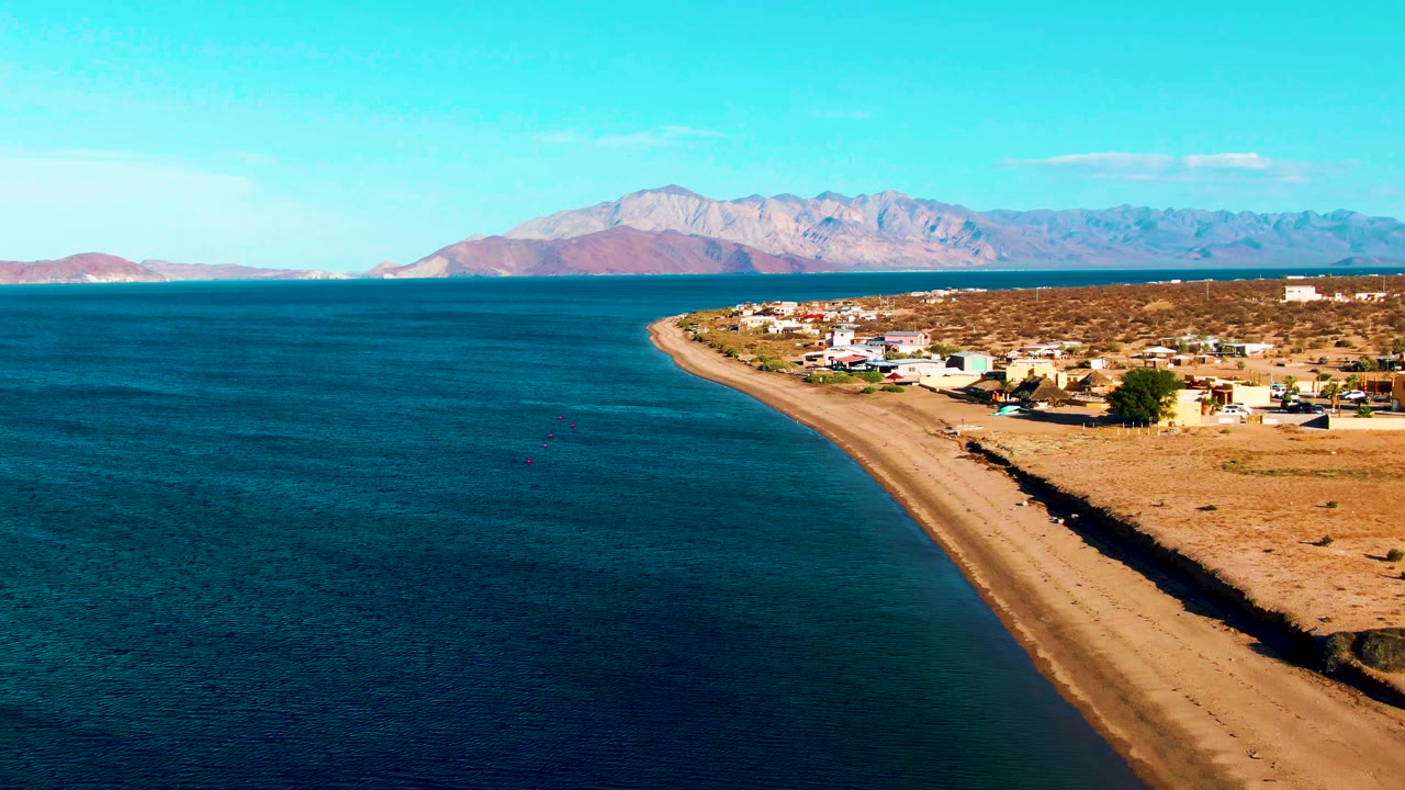 Opening aerial frame over Bahia De Los Angeles showing blue ocean beside a narrow sandy beach, low village buildings on dry land, distant mountains, clear sky, and no visible people.