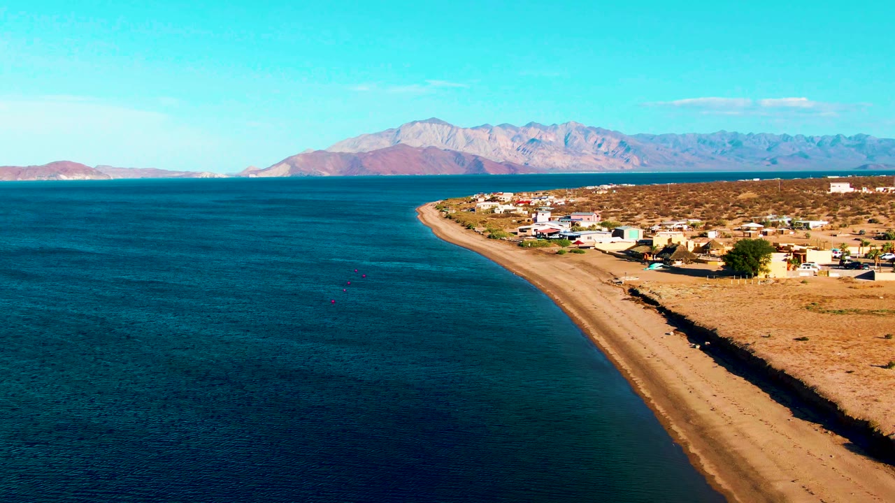 Aerial frame at 0:01 over Bahia De Los Angeles with the shoreline curving through the center, calm blue water on the left, dry village lots on the right, distant mountains, and no visible people.