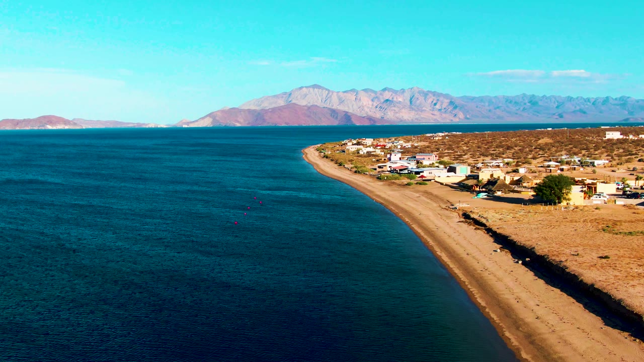 Frame at 0:02 shows Bahia De Los Angeles from above with darker blue ocean filling the foreground, a tan beach tracing the coast, scattered low buildings, distant mountains, and no visible people.