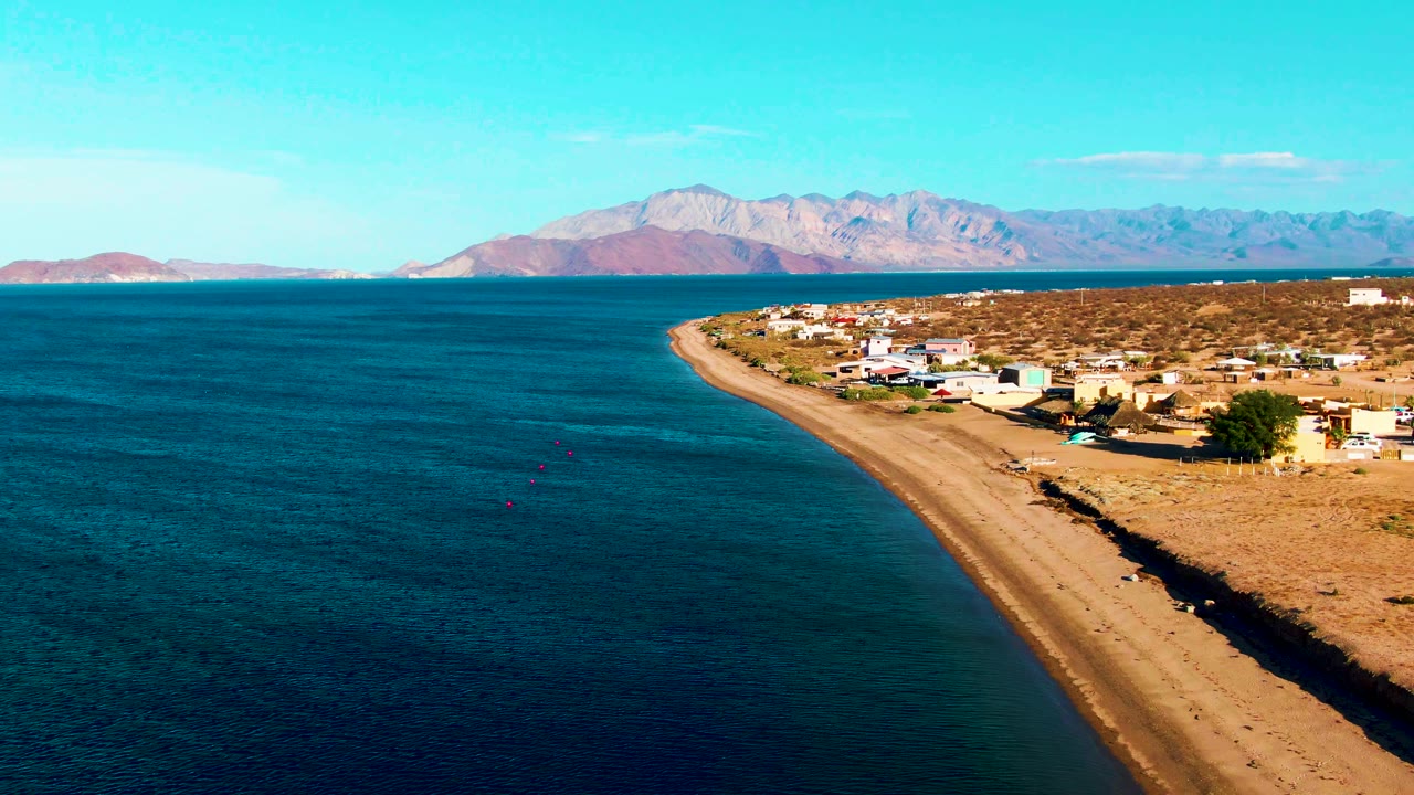 Wide aerial of ocean and sandy beach beside a sparse coastal village.
