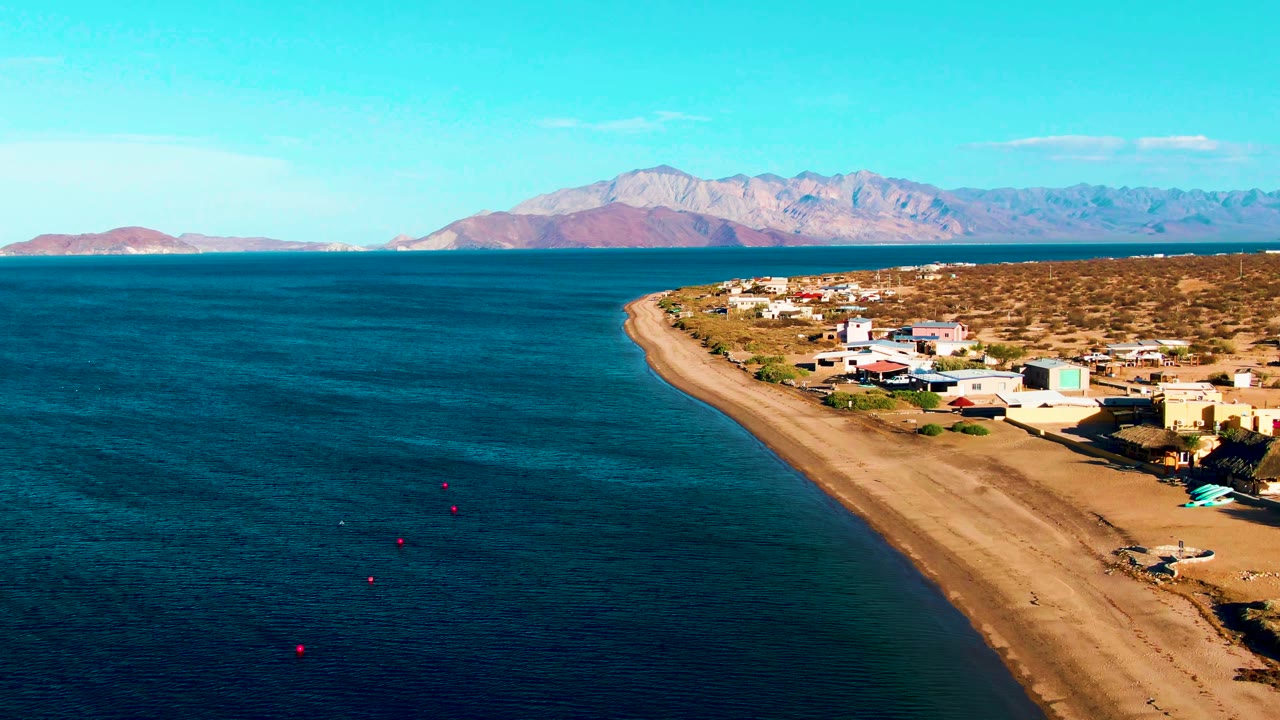 At 0:02, the drone view shows the sandy coast running diagonally, beachside structures clustered near the right edge, small buoys offshore, a pale mountain range across the horizon, and no visible people.