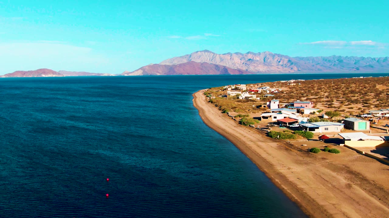 Aerial frame over Bahia De Los Angeles, Mexico, with blue water on the left, a sandy beach curving past small coastal buildings, distant mountains, and no visible people.