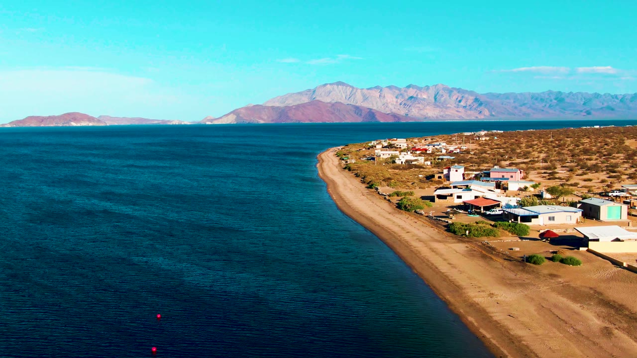 Elevated coastal view with ocean, sandy beach, low buildings, and mountains across the water.