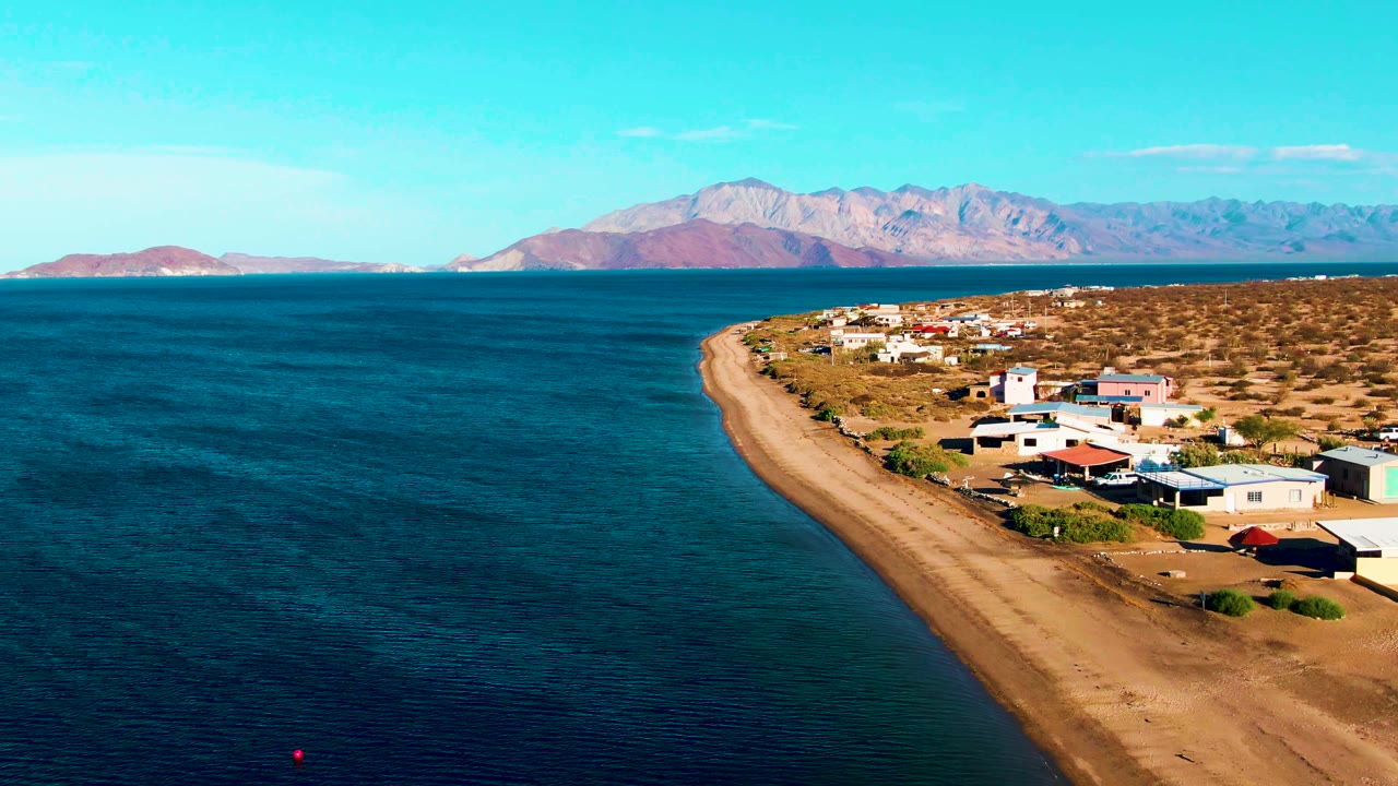 Aerial view along Bahia De Los Angeles with dark blue water in the foreground, a tan beach strip, small homes and structures, mountains across the bay, and no visible people.