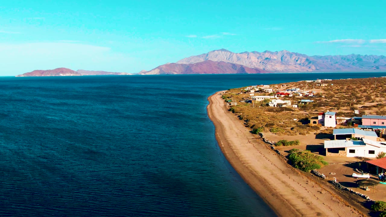 Aerial frame from Bahia De Los Angeles showing calm blue water, a curving sandy beach, scattered village buildings, distant mountains, bright sky, and no visible people.