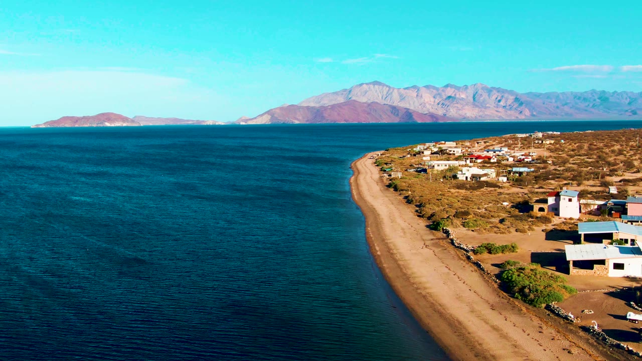Aerial view in Bahia De Los Angeles with the sandy shore running diagonally through frame, low coastal buildings at right, calm water at left, distant mountains, and no visible people.