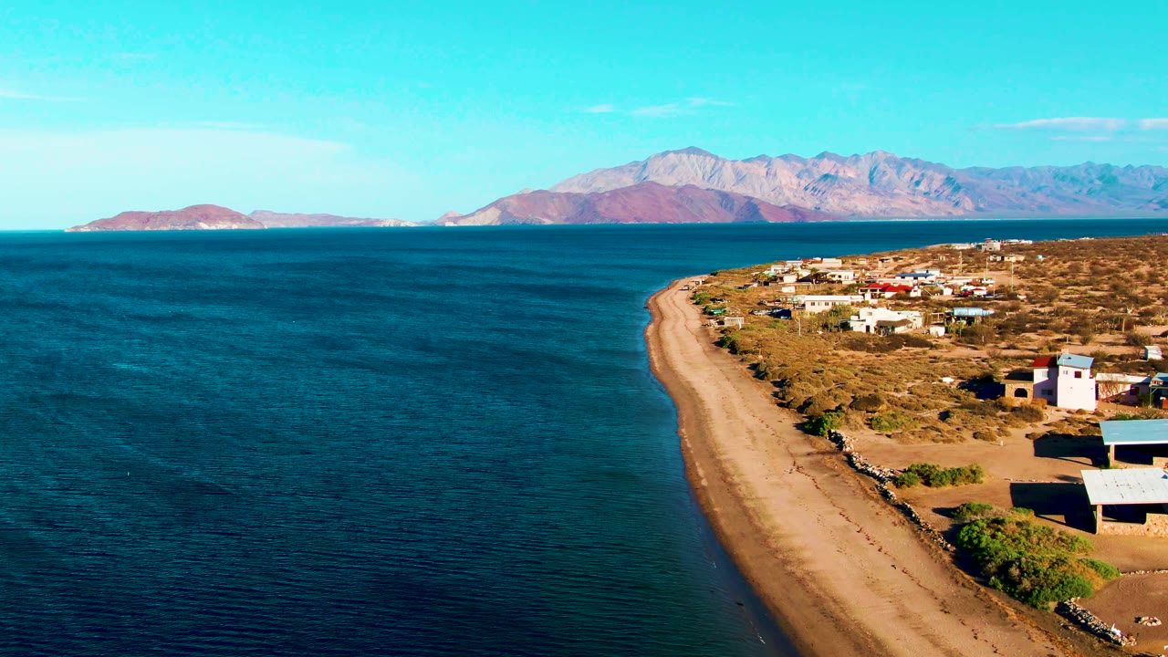 Bahia De Los Angeles aerial frame showing calm dark-blue water, tan beach, scattered houses and small structures, dry terrain, pale mountains across the bay, and no visible people.