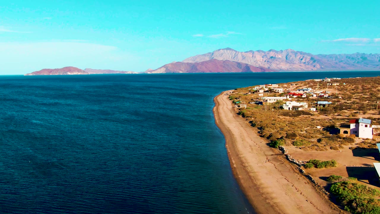 Wide aerial coastline with ocean, beach, buildings, and distant mountains.