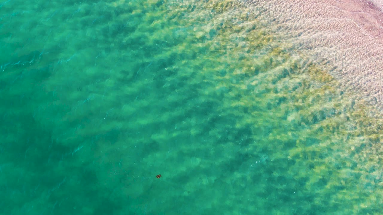 Aerial frame at Bahia De Los Angeles showing clear green-blue water over sand, faint wave lines near the upper right shore, and no visible people.