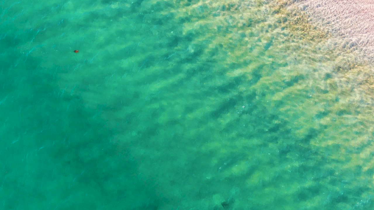 Overhead view of clear turquoise water in Bahia De Los Angeles, with soft wave bands, pale shoreline texture on the right, a small red buoy near the upper left, and no visible people.
