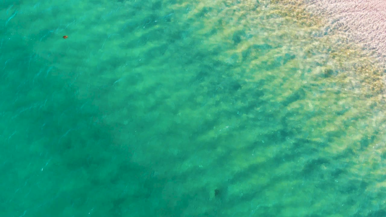 Aerial view over Bahia De Los Angeles water, with diagonal pale wave streaks crossing the turquoise shallows, a tiny red buoy left of center, and no visible people.