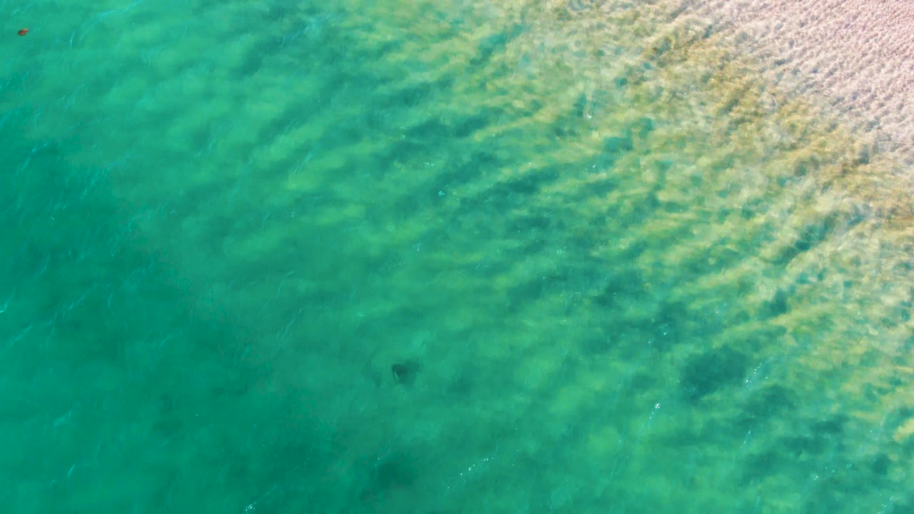 Clear green-blue water fills the frame in Bahia De Los Angeles, with the sandy shallows still visible at upper right, ripples crossing diagonally, a dark submerged object near the lower middle, and no visible people.