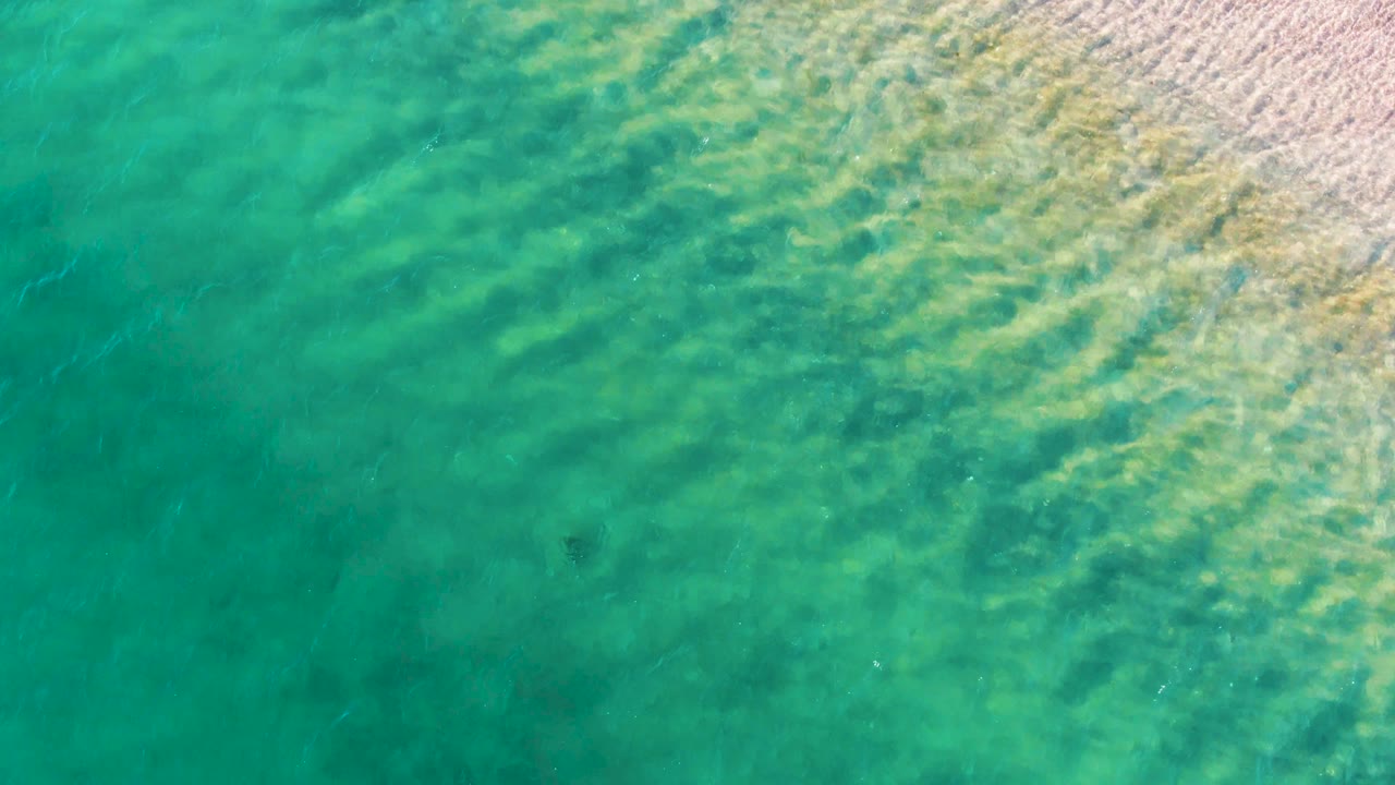 Turquoise ocean water in Bahia De Los Angeles shows brighter diagonal ripple bands, pale sand along the upper right edge, faint underwater patches, a small dark submerged object left of center, and no visible people.