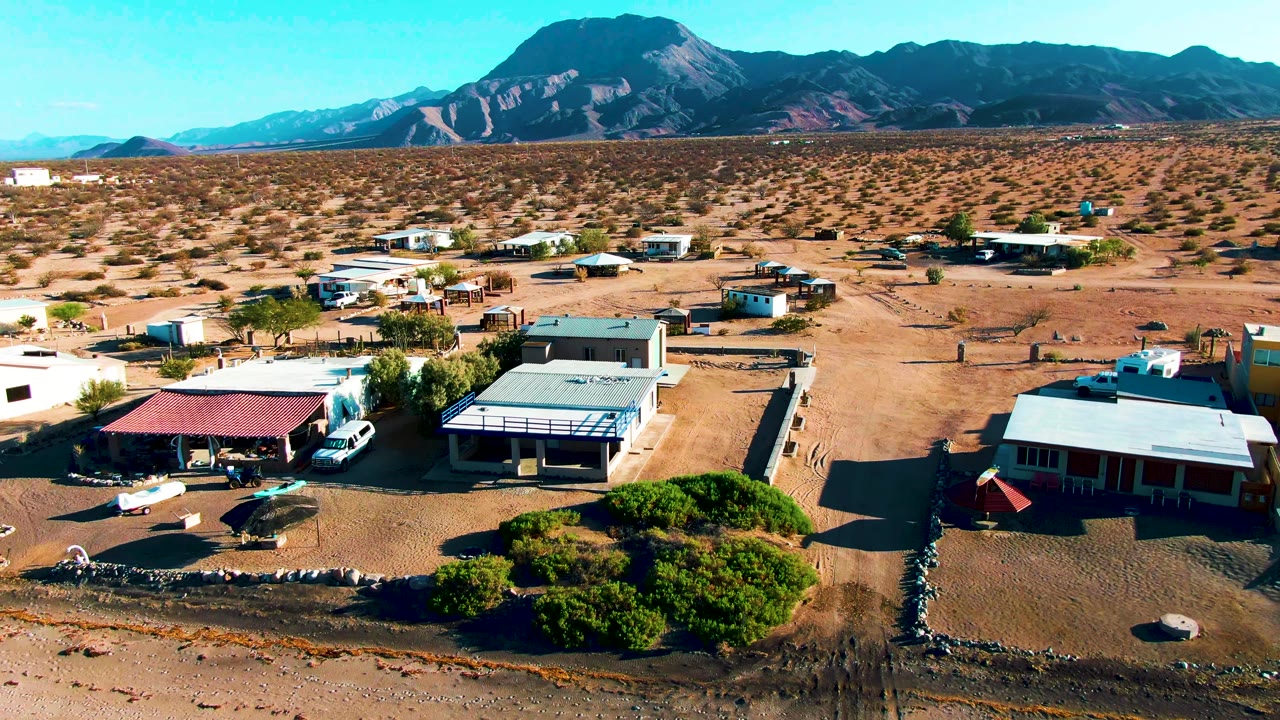 Aerial frame over Bahia De Los Angeles showing low desert buildings, parked vehicles, sandy lots, green scrub in front, mountain ridges in back, and no visible people.