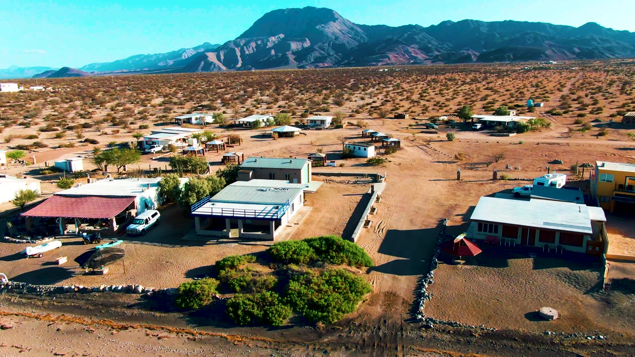 Drone perspective over Bahia De Los Angeles with a red-roofed shade structure, white vehicle, flat-roofed buildings, open sandy ground, distant mountains, and no visible people.