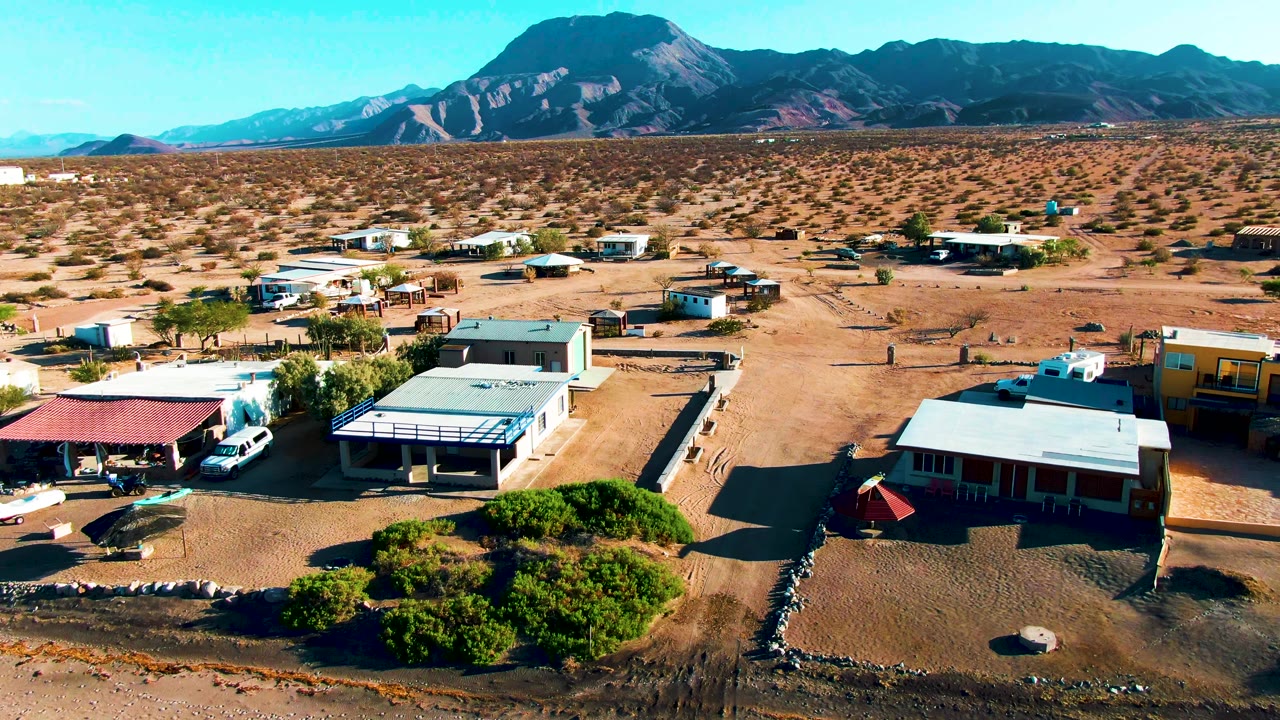 Aerial Bahia De Los Angeles frame showing a yellow building at right, low homes or structures, parked vehicles, open desert scrub, distant ridges, and no visible people.