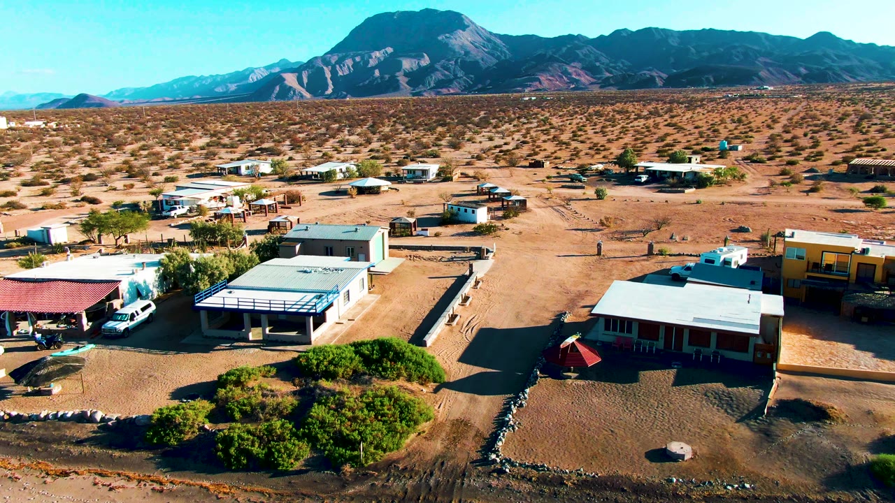 Wide drone frame over low desert buildings, vehicles, shrubs, and mountain horizon.