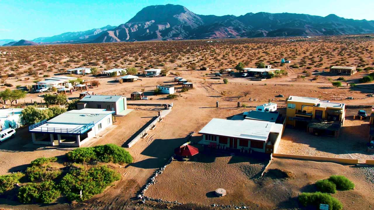 Aerial view over Bahia De Los Angeles mobile homes and sandy lots, with sparse shrubs, a clear sky, distant mountains, and no visible people.