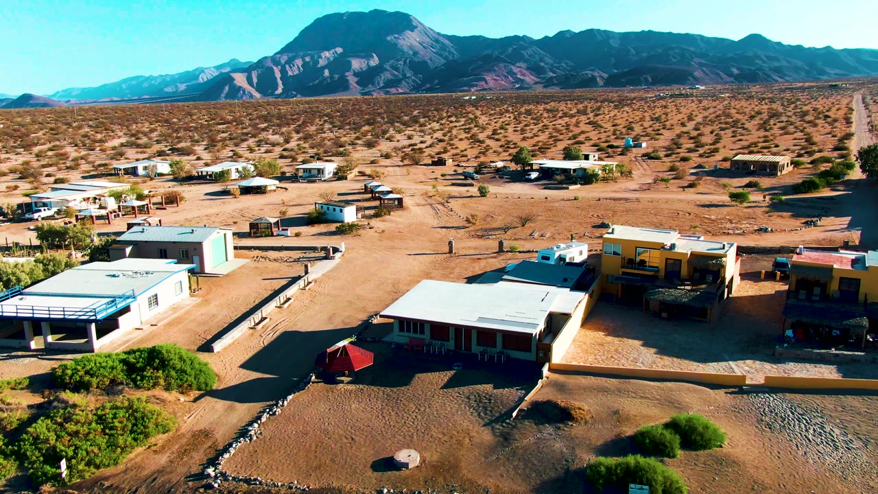 Aerial frame of Bahia De Los Angeles homes and sandy roads, with scattered shrubs, flat-roofed structures, a distant road line, mountains, and no visible people.