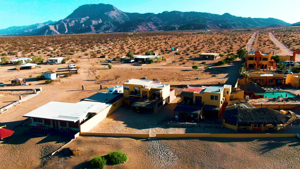 Aerial frame of Bahia De Los Angeles showing low desert buildings, a small pool, sandy lots, a straight road, sparse scrub, mountains, blue sky, and no visible people.