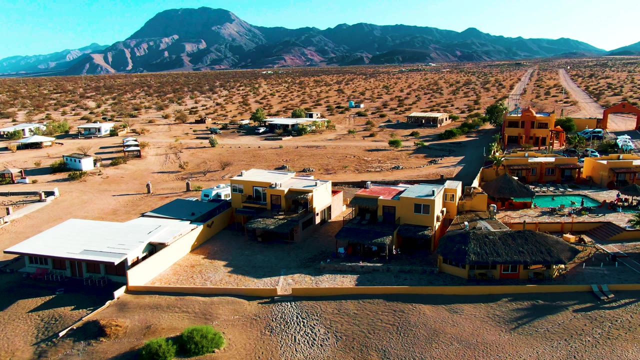 Aerial view in Bahia De Los Angeles with yellow and white buildings around a small pool, open desert lots, a road leading toward distant mountains, and no visible people.