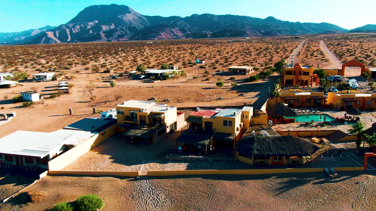 High aerial frame of Bahia De Los Angeles with foreground roofs, a pool area, parked vehicles, desert scrub, a long road, mountain ridges, blue sky, and no visible people.