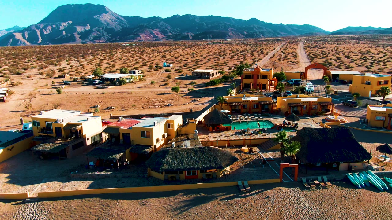 Aerial view over a Bahia De Los Angeles desert resort, with yellow buildings, palapa roofs, a turquoise pool, parked vehicles, sparse scrub, and mountains beyond.