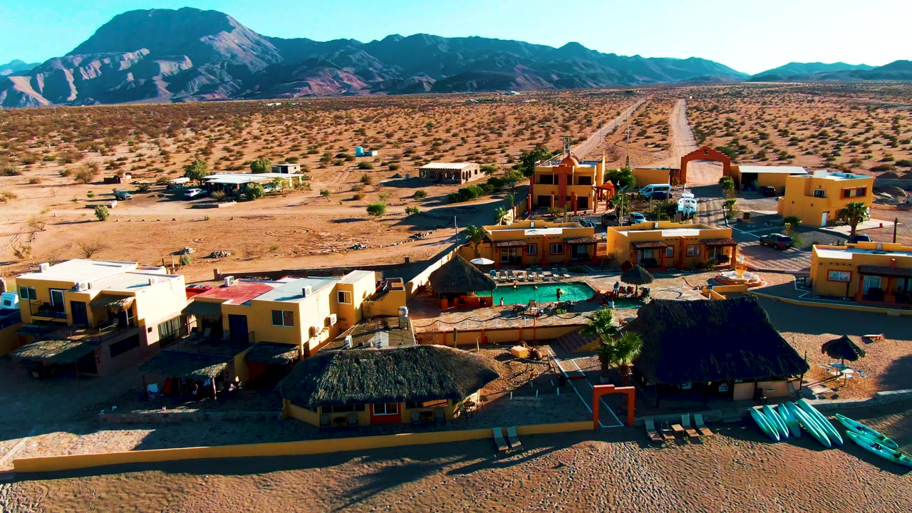 Wide drone view of resort buildings, pool, kayaks, desert road, and mountains.