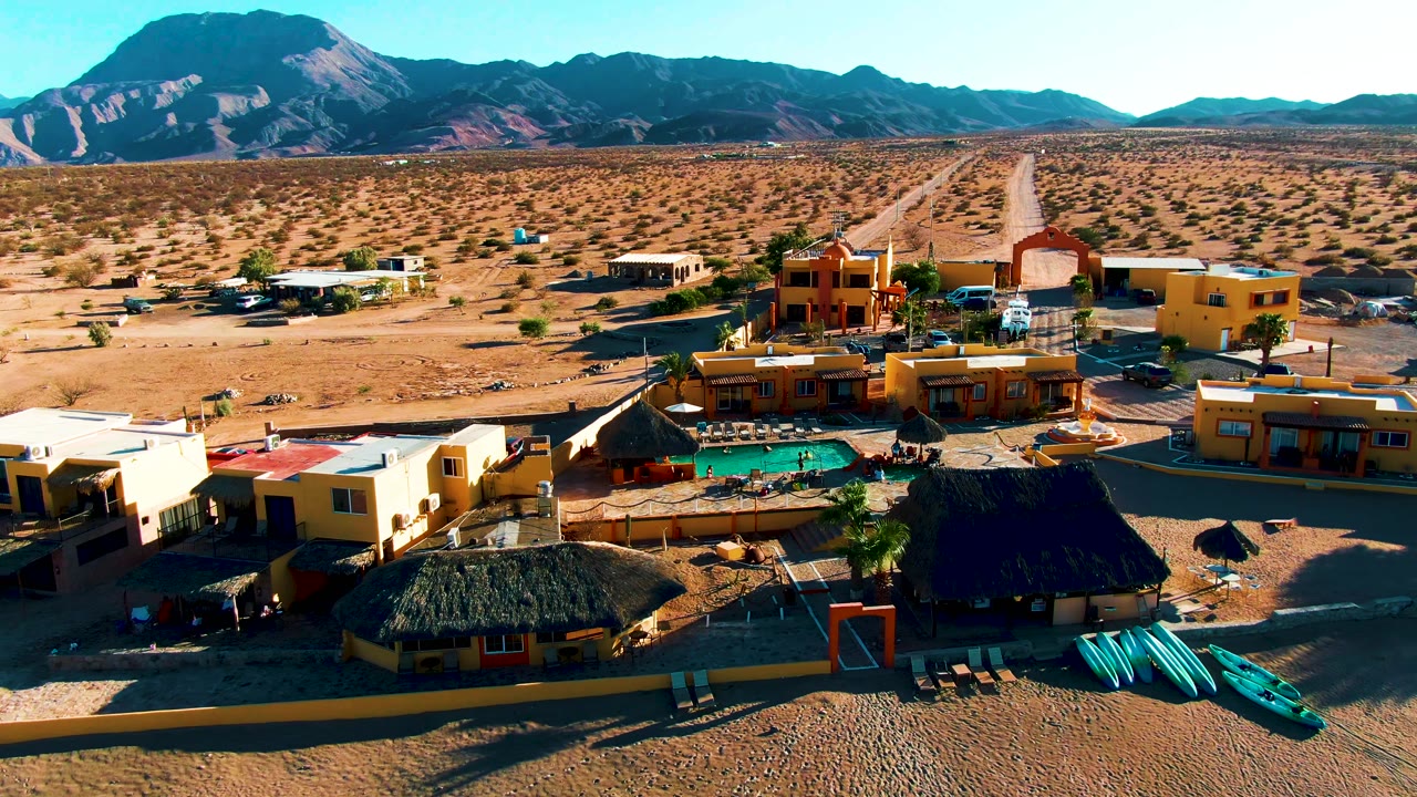 A Bahia De Los Angeles resort compound fills the lower frame, with palapa roofs, a bright pool, lounge chairs, vehicles, dry open ground, a long road, and dark mountains behind it.