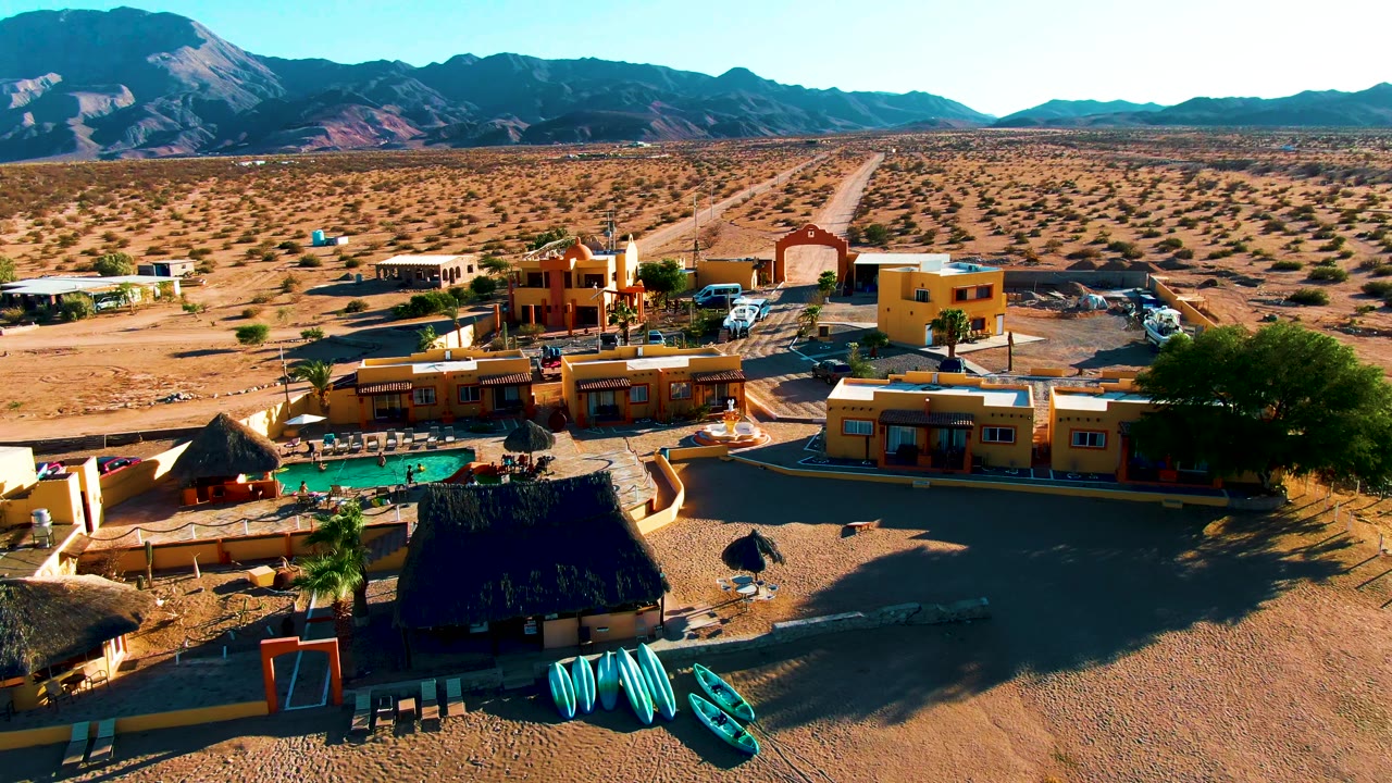 Aerial frame over a Bahia De Los Angeles desert resort with yellow buildings, a turquoise pool, kayaks in the foreground, scrubland and mountains beyond, and no visible people.