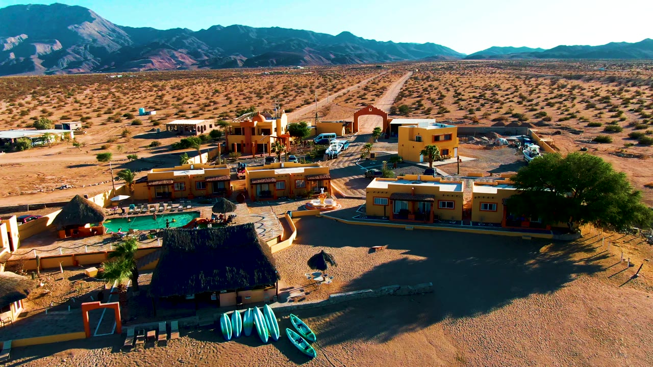 Elevated view of the Bahia De Los Angeles resort shifted slightly along the property, showing the pool at lower left, yellow buildings, parked vehicles, a straight desert road, and no visible people.
