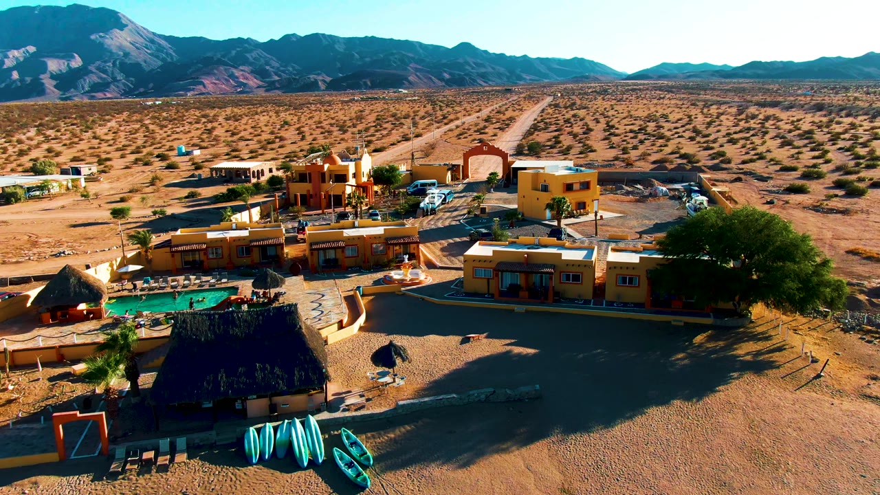 Wide aerial frame in Bahia De Los Angeles with the resort centered lower in the image, the pool and shade structures near the foreground, open scrubland behind, and no visible people.