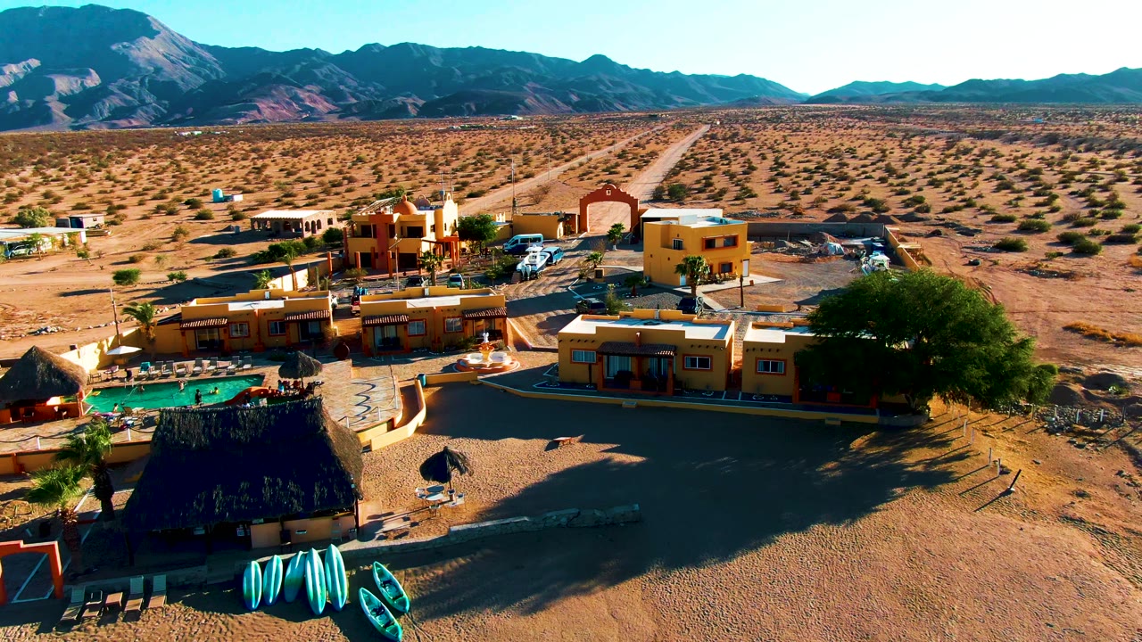 Aerial resort view with dark foreground roof, kayaks, pool, yellow buildings, desert road, and mountains.