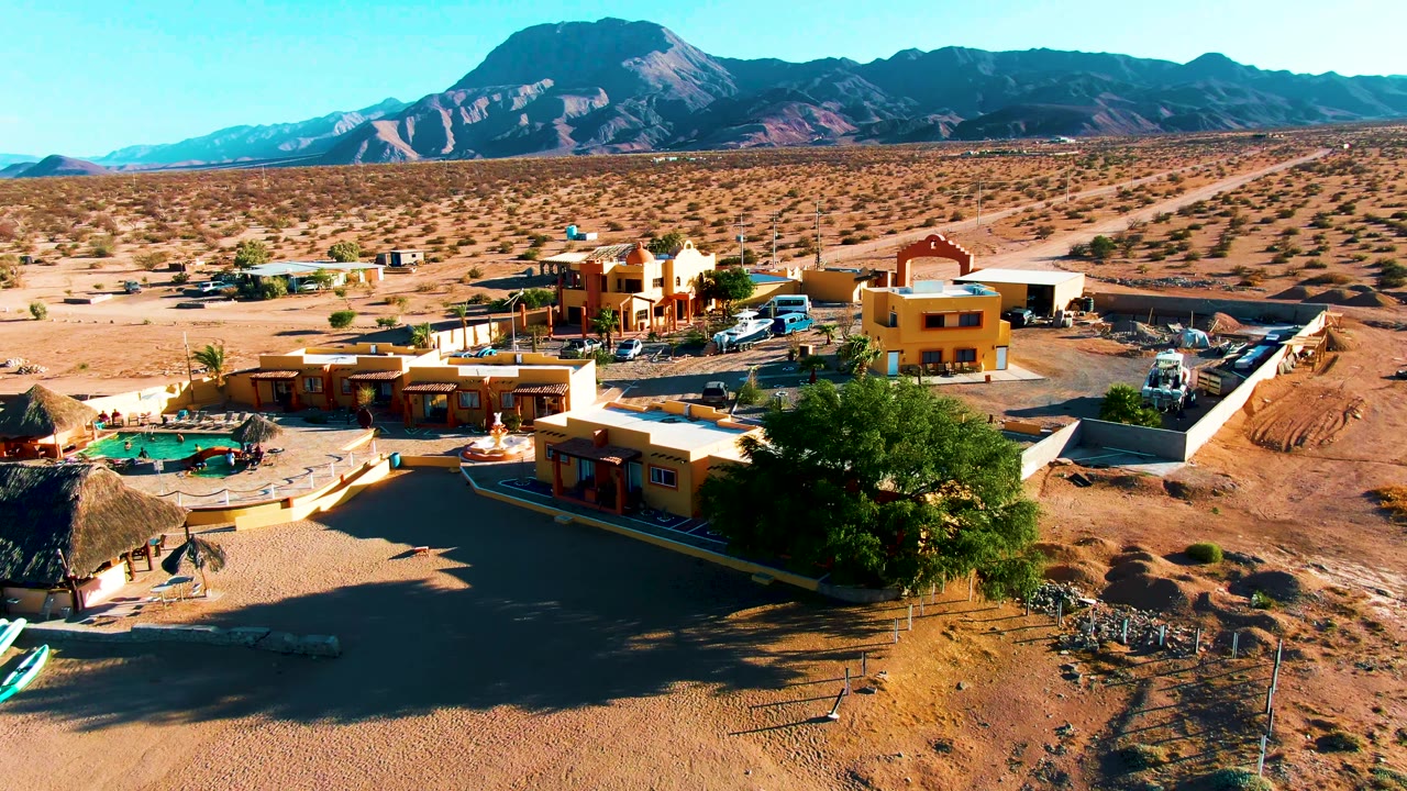 Opening aerial frame over Bahia De Los Angeles, Mexico, showing low desert buildings, a green pool, a large foreground tree, scattered vehicles, sandy roads, distant mountains, and no visible people.