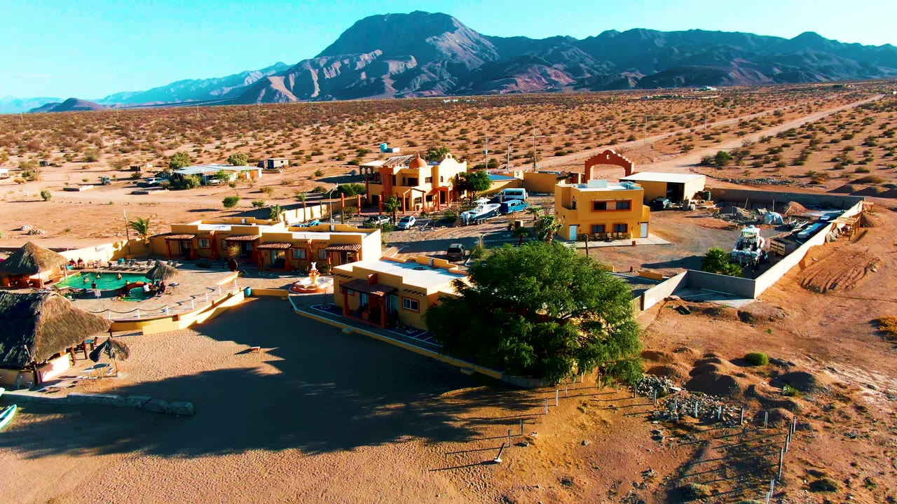 Wide drone view of sandy buildings, pool, vehicles, tree shade, and mountain ridge.