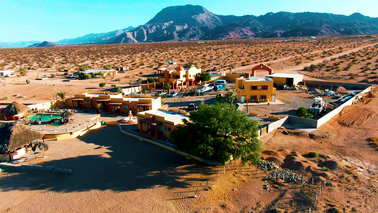 At 0:02, the Bahia De Los Angeles frame keeps the pool left of center, yellow and earth-toned buildings to the right, vehicles near the structures, open desert behind them, and no visible people.