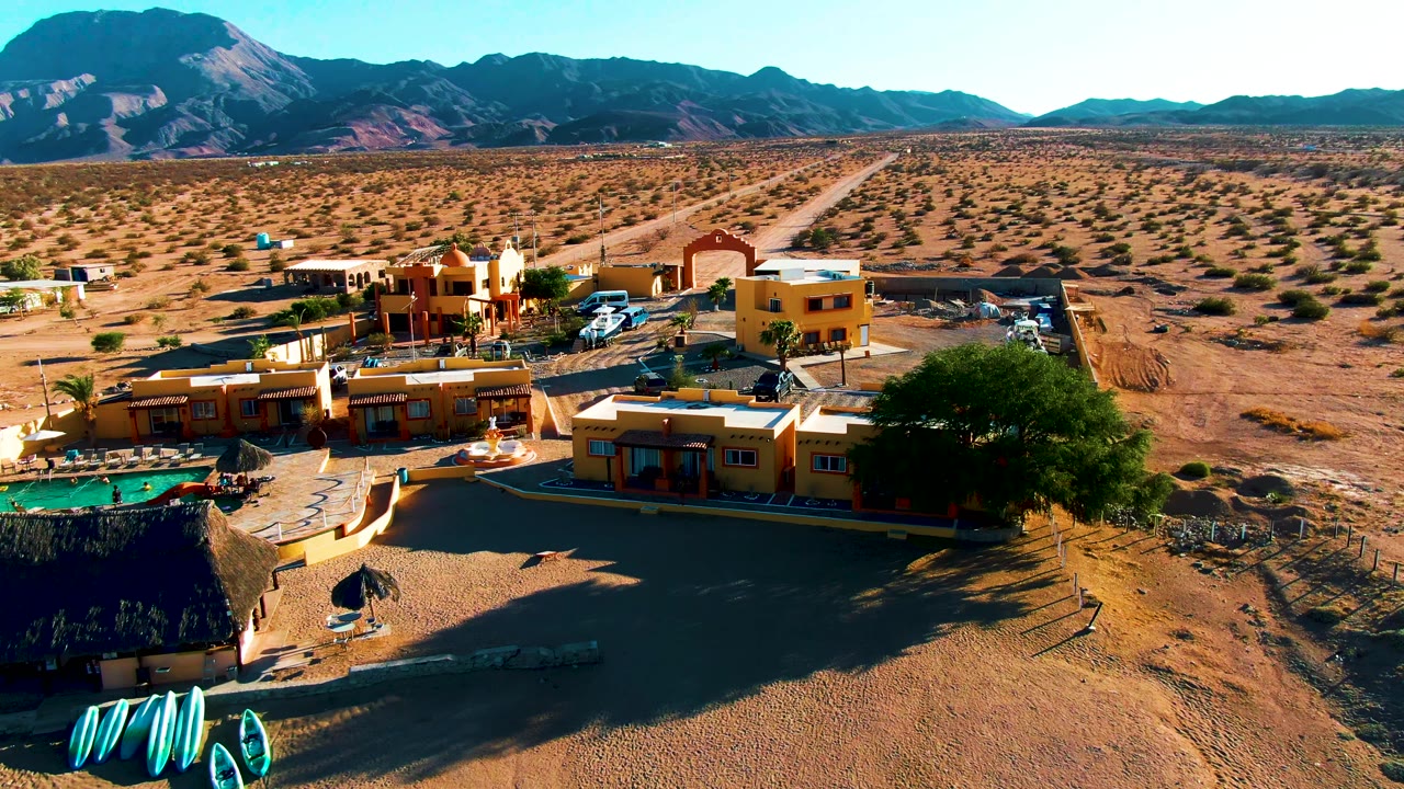 A high aerial frame over Bahia De Los Angeles shows yellow buildings, a pool area, parked vehicles, sandy desert, distant mountains, and no visible people.