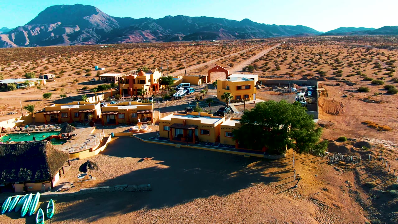 A Bahia De Los Angeles aerial view shows low earth-toned structures, parked cars, a small pool, broad sandy ground, sparse shrubs, mountain ridges, and no visible people.