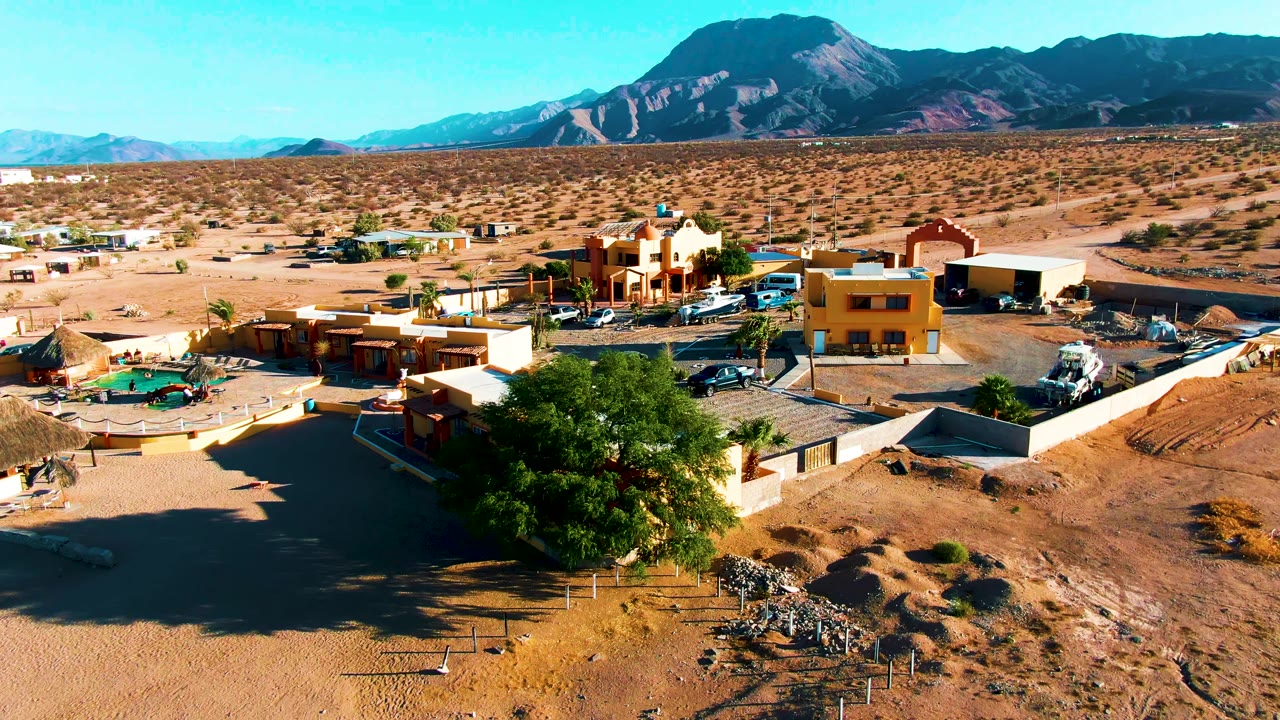 Wide aerial frame over Bahia De Los Angeles showing sandy lots, low buildings, a large green tree, parked vehicles, distant mountains, clear blue sky, and no visible people.