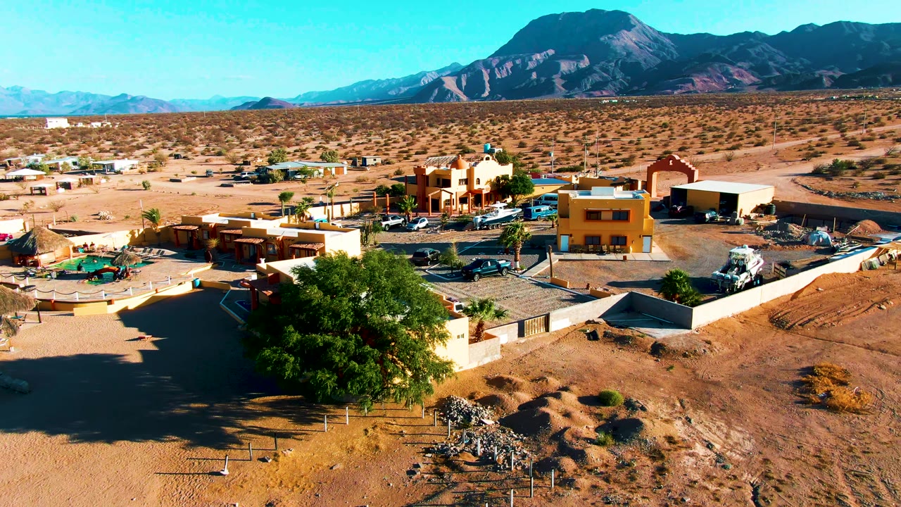 Wide drone frame over Bahia De Los Angeles with sandy terrain in front, a cluster of low buildings and parked vehicles in the midground, a broad mountain range, and no visible people.