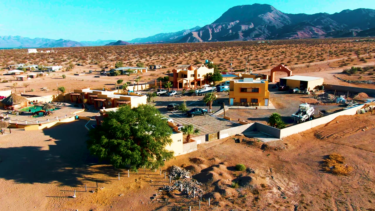 Drone view of low buildings, dirt lots, green tree, and mountains