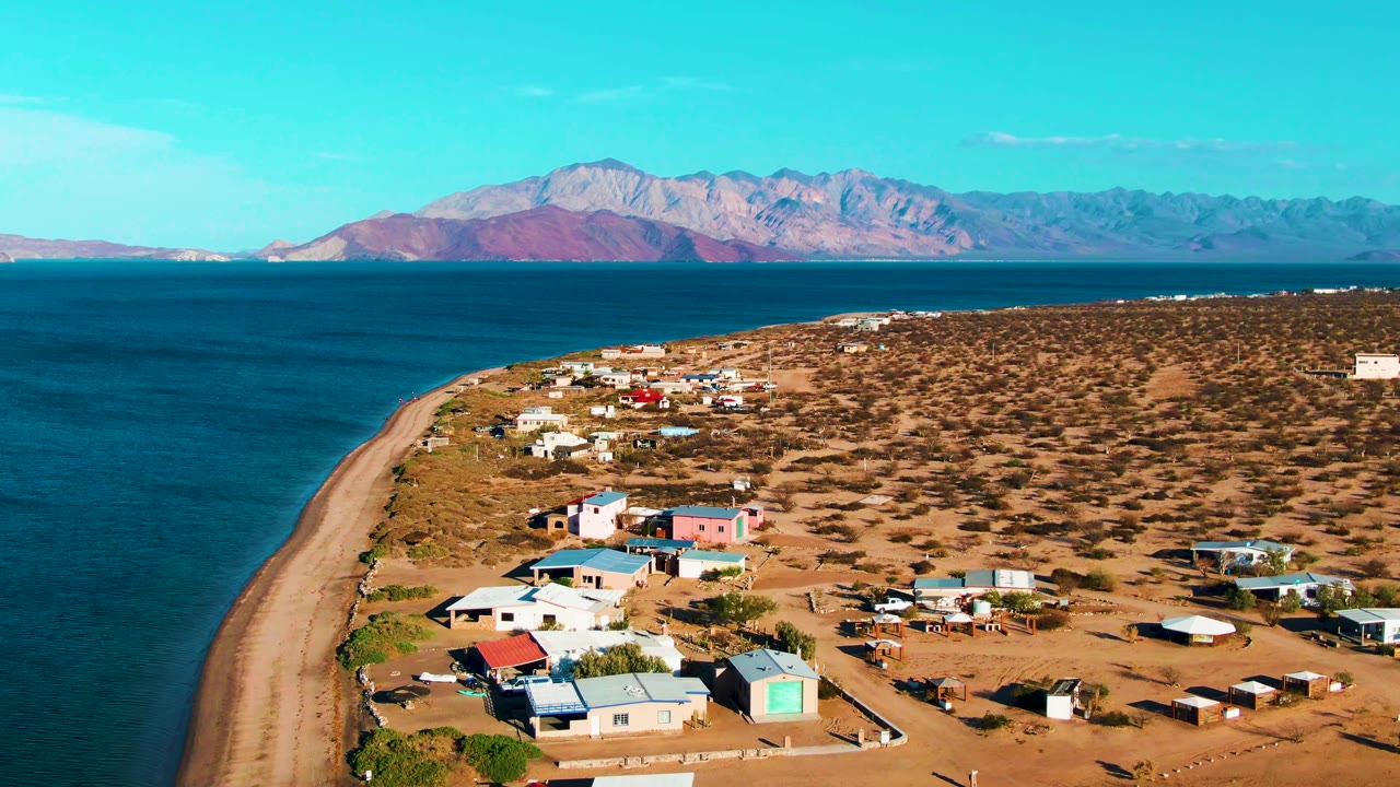 Opening aerial frame over Bahia De Los Angeles shows a curving sandy beach, scattered low buildings, dry scrubland, blue ocean, distant mountains, and no visible people.