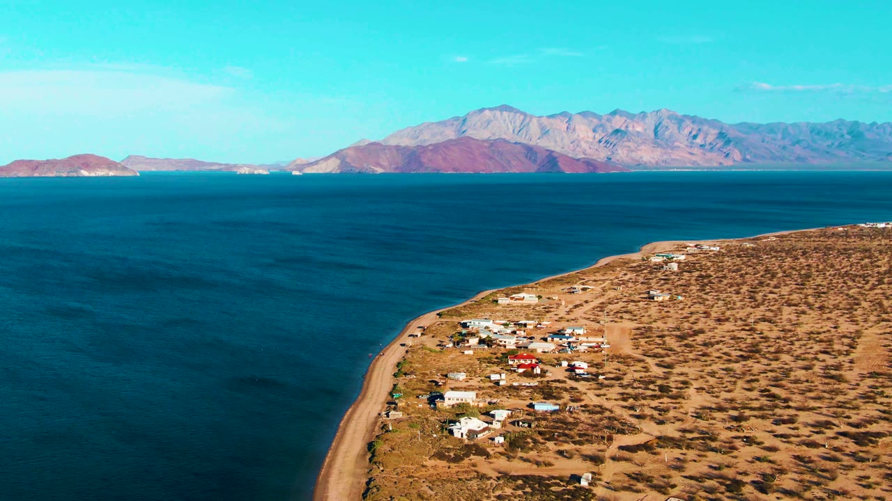 Opening aerial frame over Bahia De Los Angeles, with a sandy shoreline and small houses below, blue ocean across the center, distant mountains on the horizon, and no visible people.