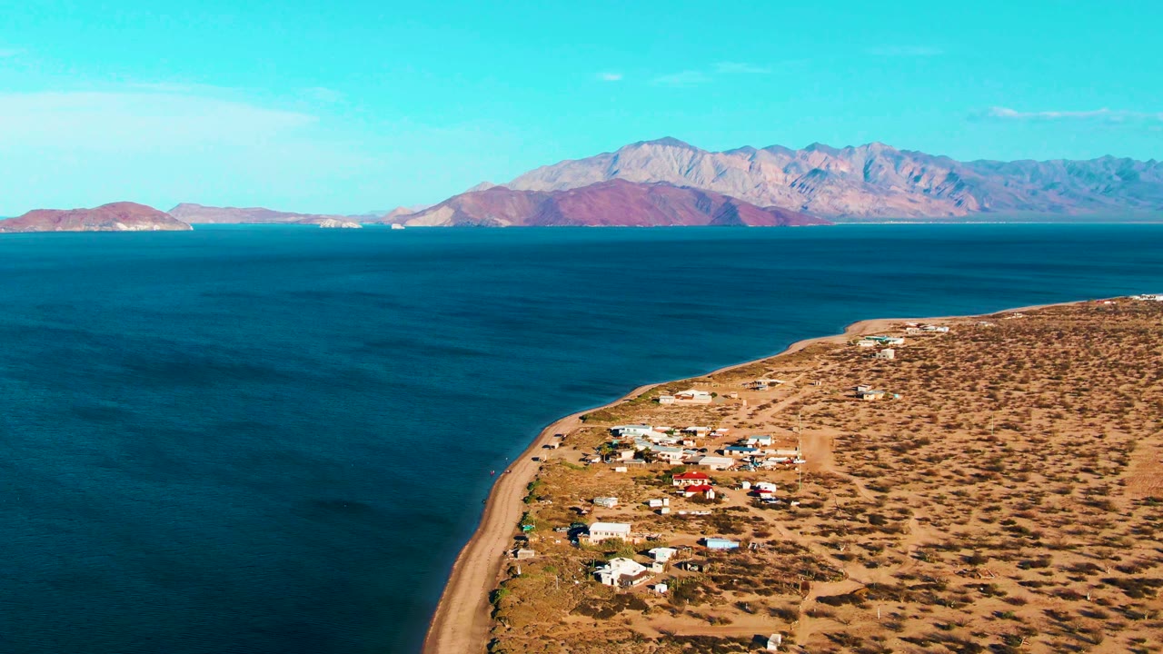 Aerial view one second into the Bahia De Los Angeles clip, showing the curving sandy shore, scattered homes, dry scrubland, blue water, mountain ridge, and no visible people.